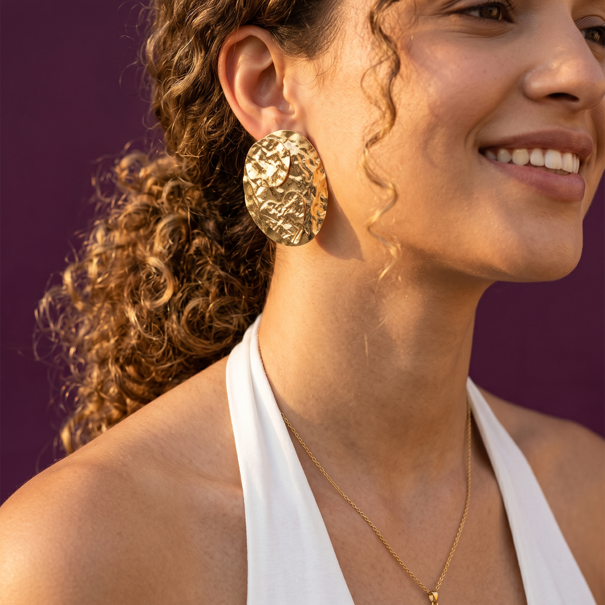 A close-up lifestyle shot of a woman wearing a chunky, textured gold earring and a delicate gold necklace, set against a solid deep purple background.