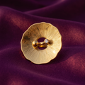 A gold-toned flower-shaped finger with a textured sunburst pattern and a small clear gemstone, displayed on a draped purple fabric background.