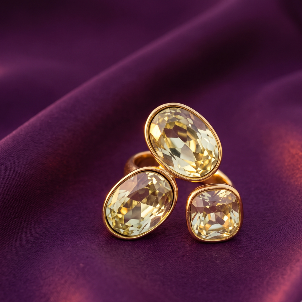 A gold ring with three oval-cut pale yellow gemstones, displayed on a draped purple fabric background.