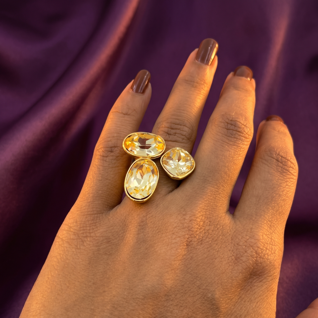 A close-up of a hand wearing a gold ring with three faceted pale yellow gemstones, set against a draped purple fabric background.