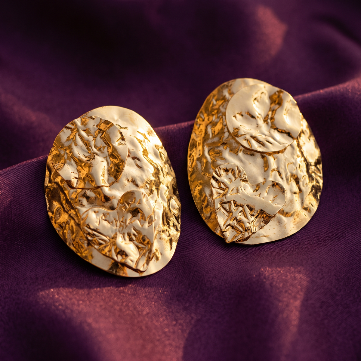 Two irregularly shaped gold earrings with a textured, hammered organic surface, displayed on a draped purple fabric background.