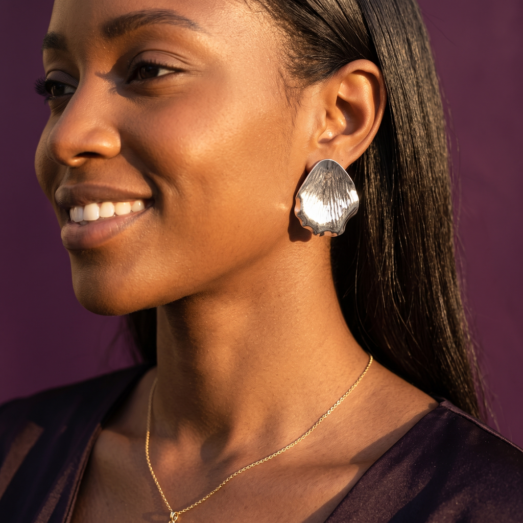 A close-up of a smiling woman wearing a large, textured silver earring and a delicate gold necklace against a solid dark purple background.