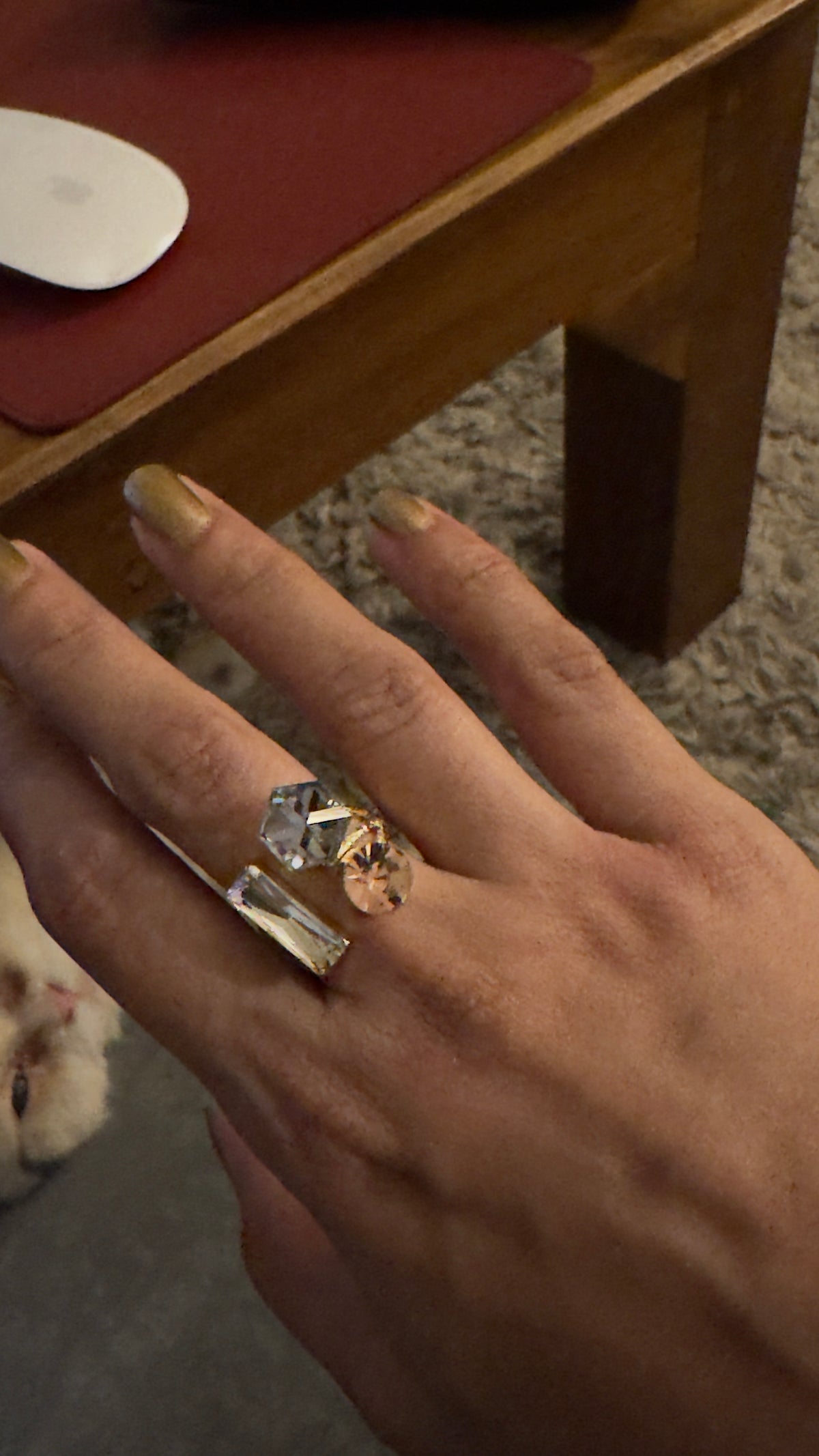 A close-up of a hand wearing a statement ring with a large clear rectangular gemstone and a smaller gold-toned geometric stone, shown in an indoor setting.