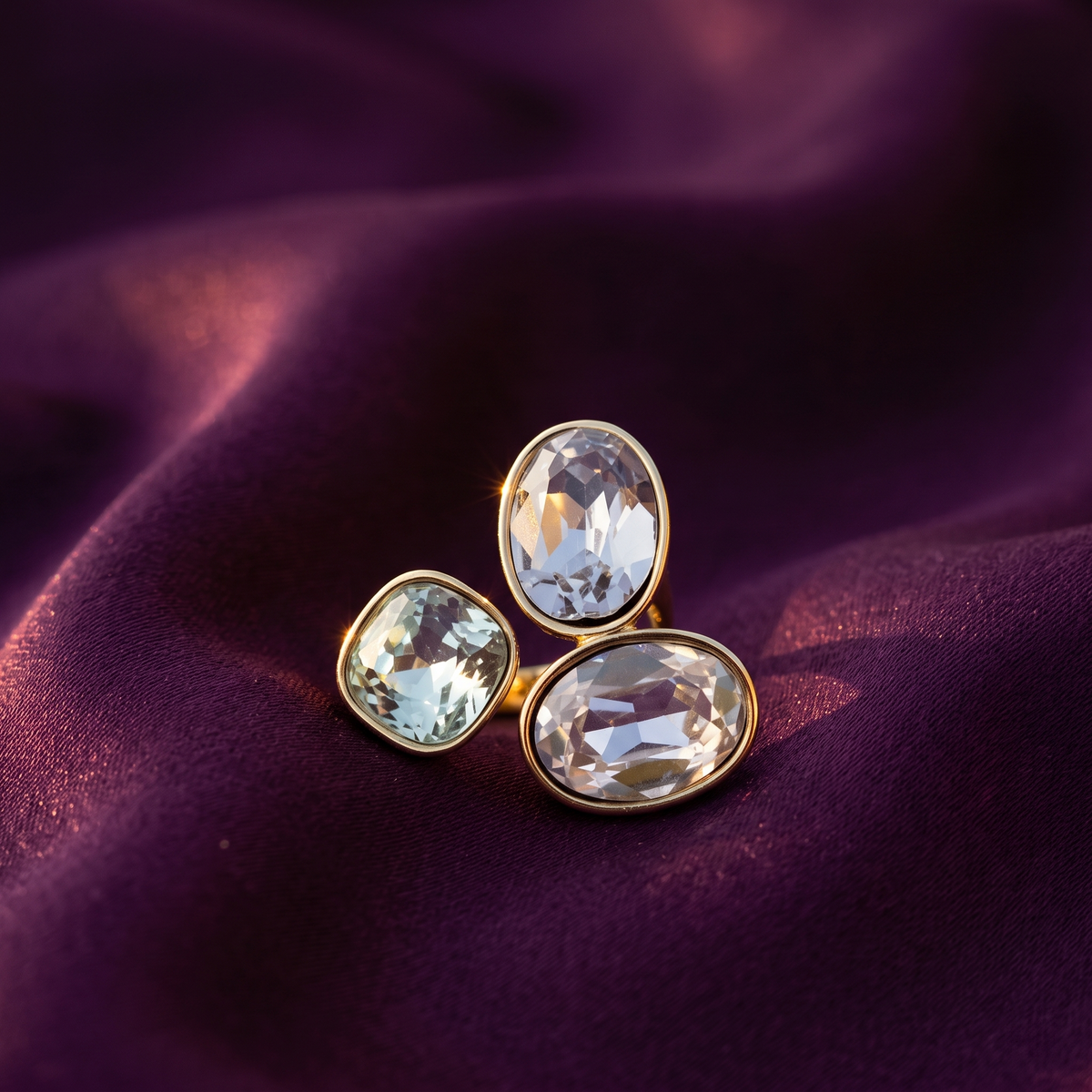 Three oval and round clear gemstone stud earrings in gold settings, arranged on draped deep purple satin fabric.
