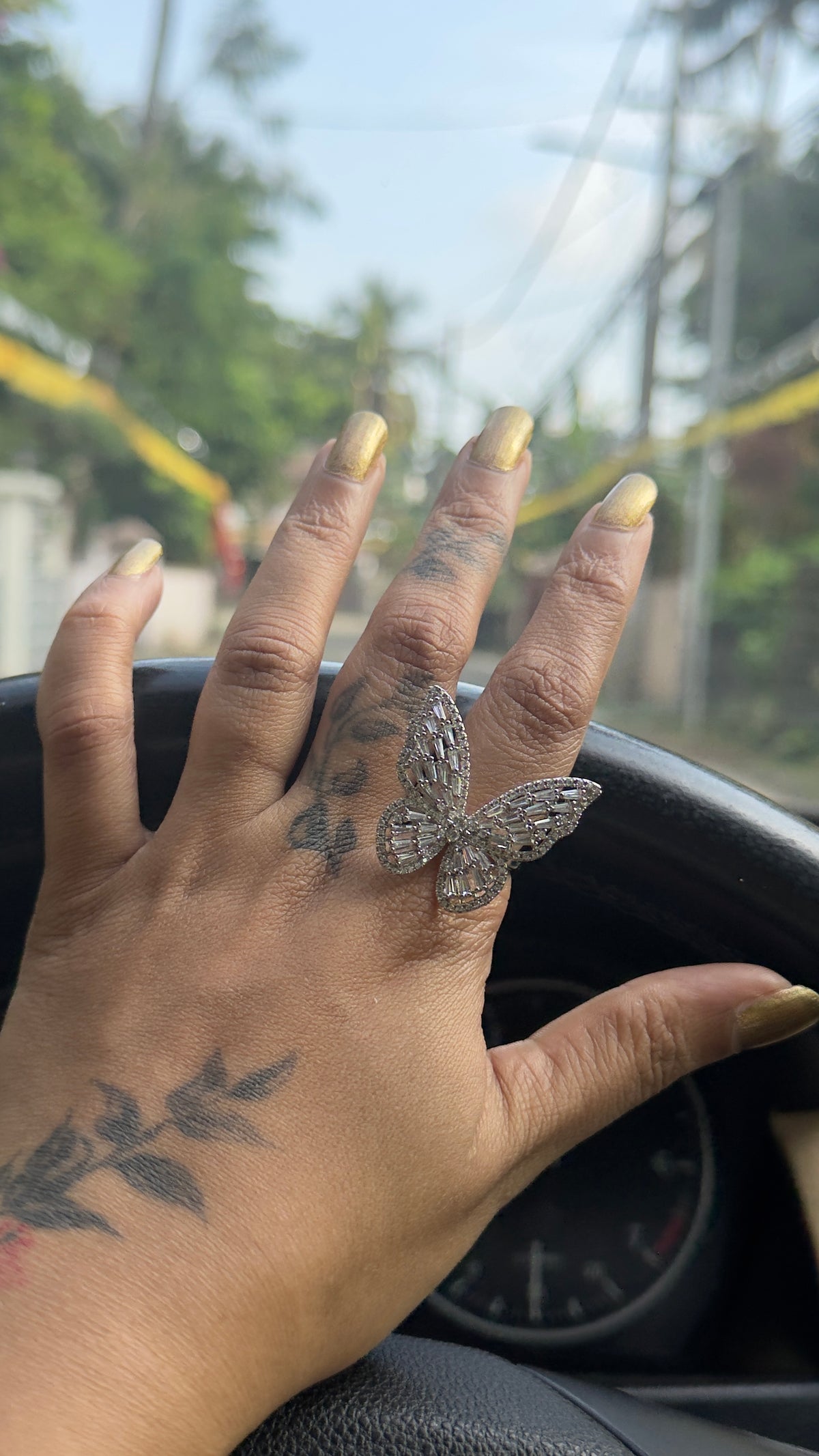 A close-up of a hand resting on a car dashboard, wearing a large silver butterfly ring on the ring finger. The hand has yellow manicured nails and black tattoos on the back and wrist.